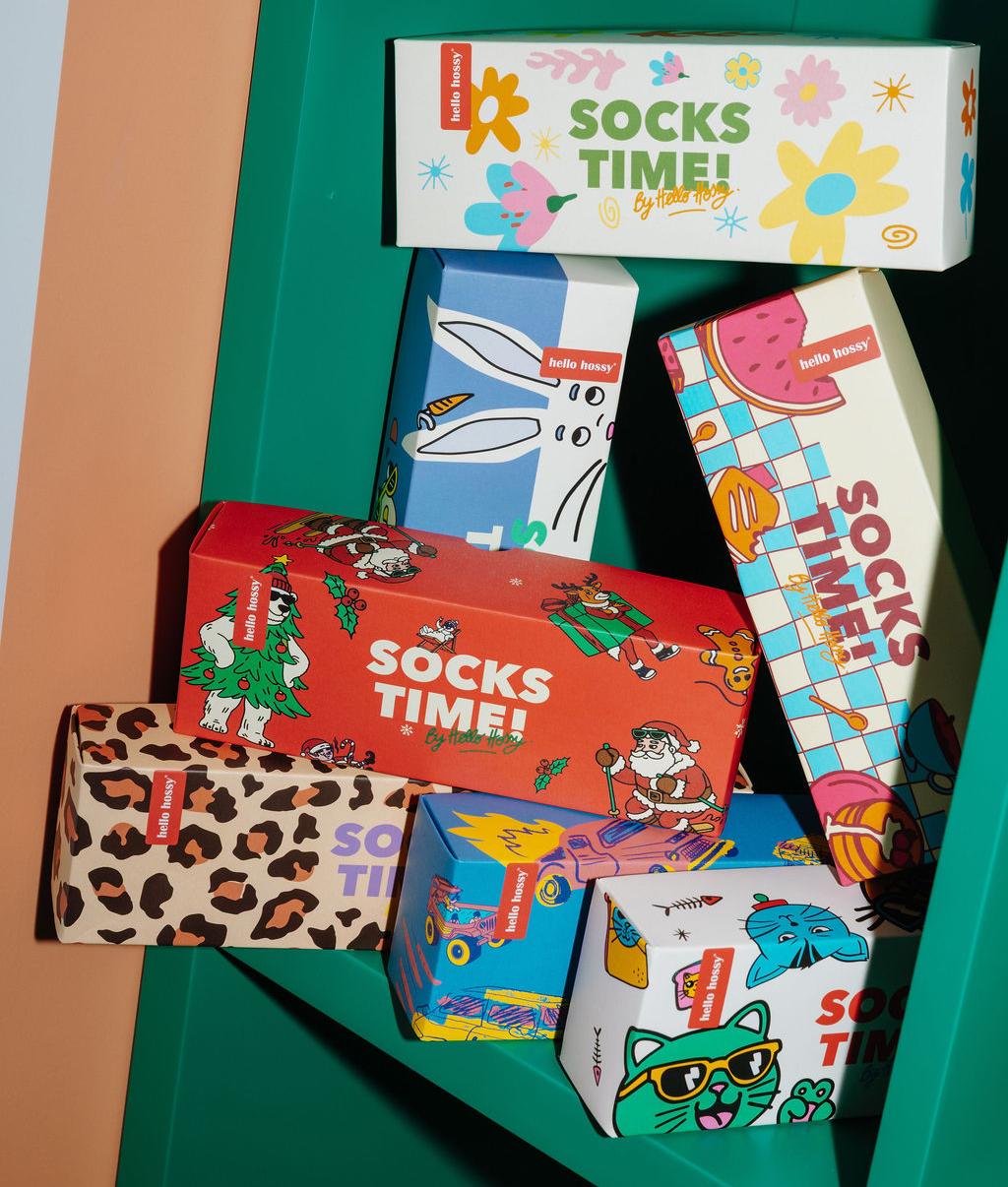 Coffrets cadeaux "Socks Time !" empilés sur une étagère verte, packagings colorés et illustrés : motifs Noël, fleurs, léopard, mascottes, voitures, chats et nourriture – Hello Hossy