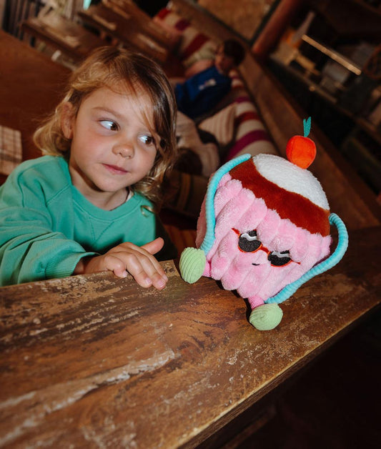 Peluche en forme de gâteau avec lunettes Cherry Cake posée sur une rampe avec un enfant la regardant - jouet original - Hello Hossy